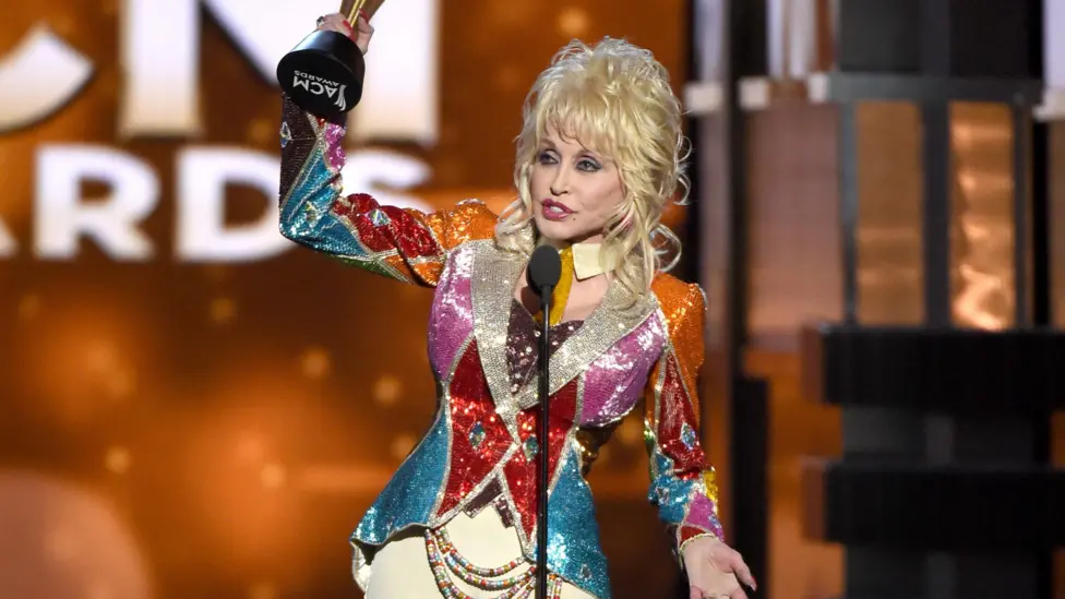Dolly holds up the award in the air. She wears a riding jacket type coloured shiny, sparkly jacket, with cream jodhpurs.