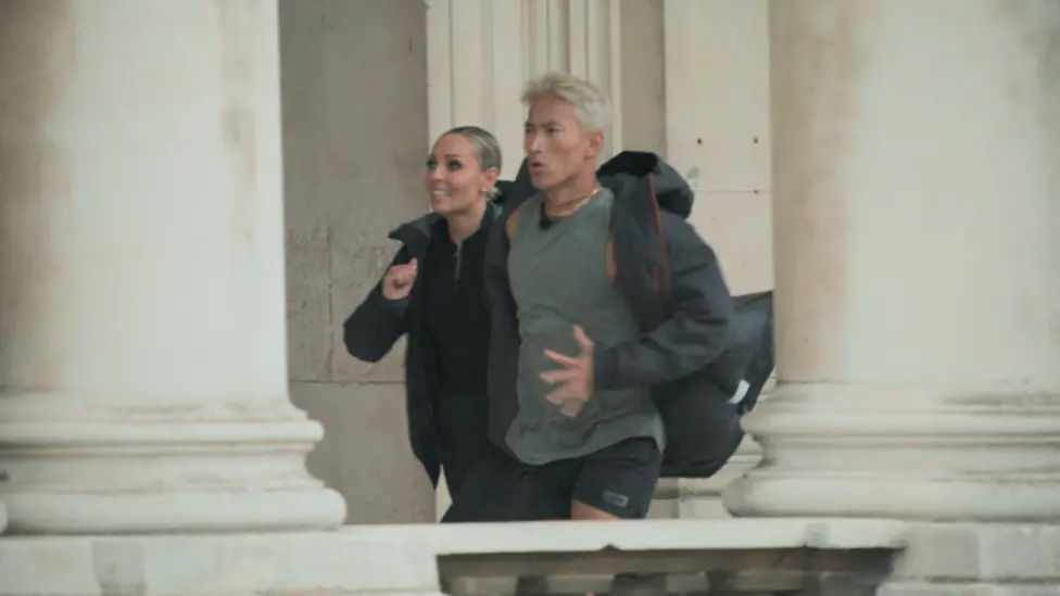 Carlos Gu and Amy Dowden pictured running side by side during an episode of the Channel 4 show, Celebrity Hunted. Carlos is wearing a grey dark coat, a light grey t-shirt and several necklaces. Amy is wearing a black top