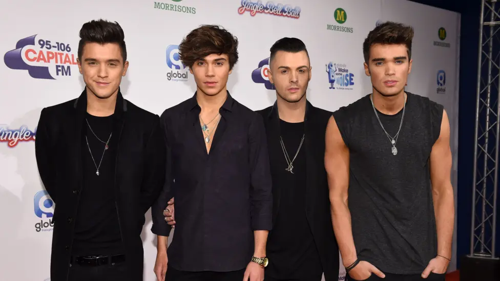 Four members of a boy band are standing side by side. They are all wearing black clothes and long necklaces. They are standing in front of a white sign.