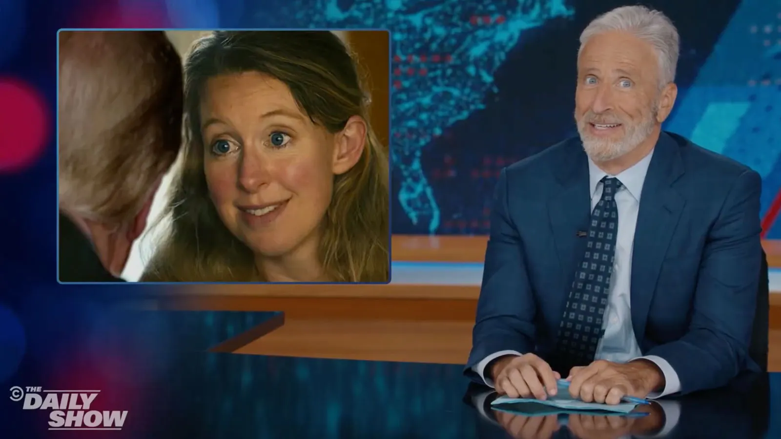 Jon Stewart speaking behind the anchor desk on The Daily Show set.