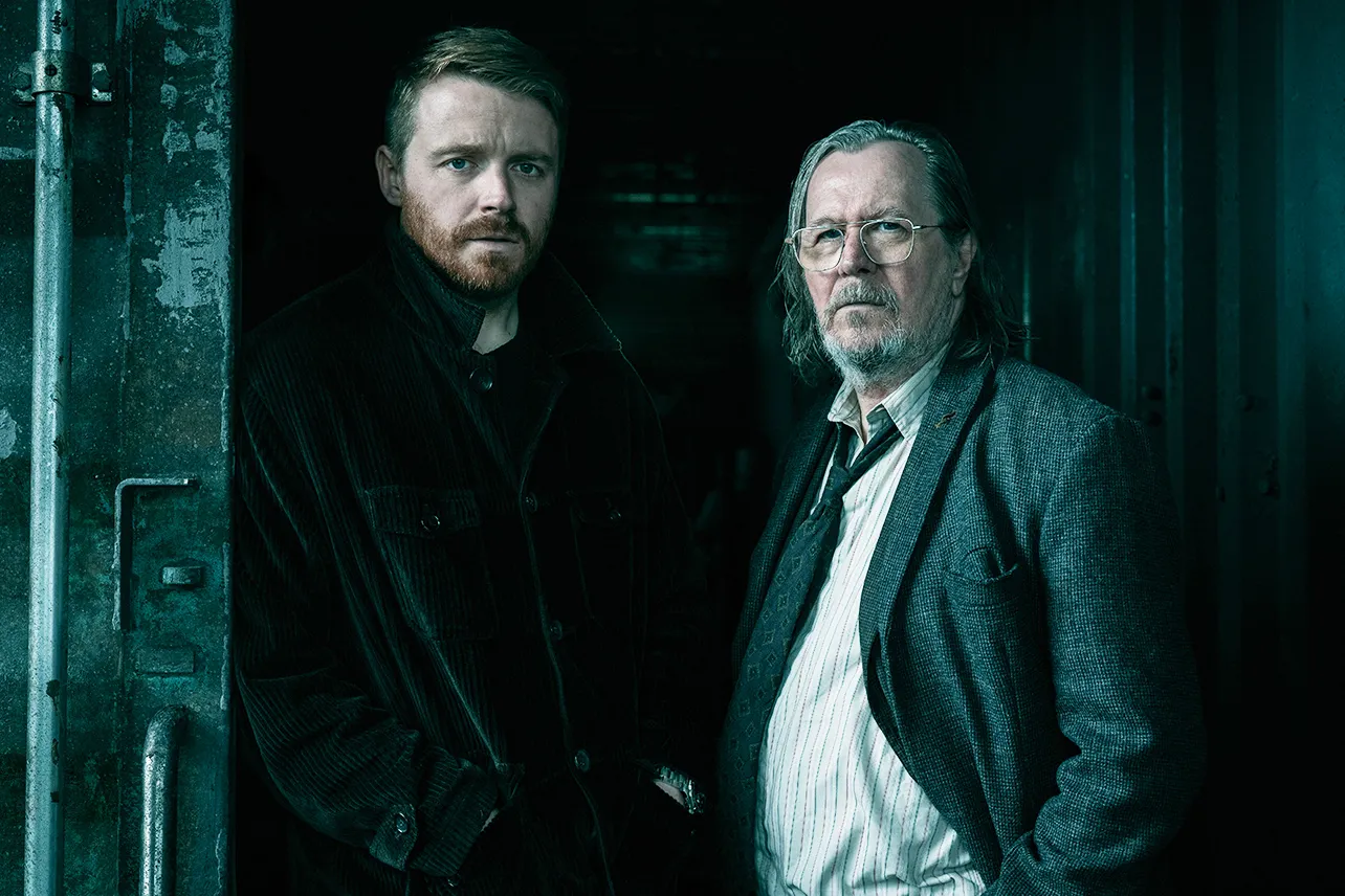 Two men (Jack Lowden and Sir Gary Oldman) standing side by side with hands in their jacket pockets and serious faces