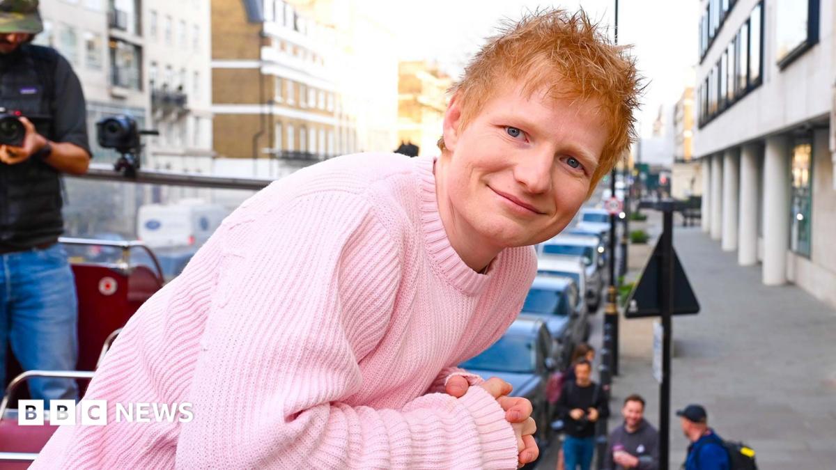Ed Sheeran smiling smiling and looking at the camera wearing a pink jumper. He is on the top deck of a bus in London