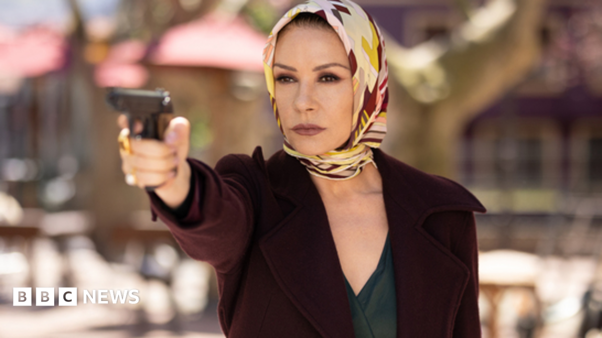 Catherine Zeta-Jones holding a gun up, wearing a scarf around her head and a maroon jacket in the Kill Jackie series.