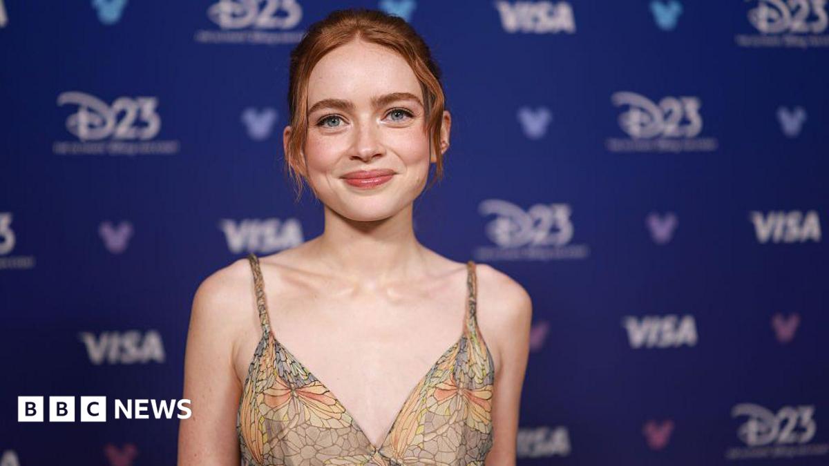 Sadie Sink smiling for the camera, she is wearing a strappy top that has a pattern on it that looks like stained-glass windows. She has auburn hair tied up and behind her is a blue advertising board for Disney's D23 fan