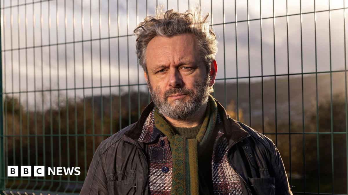 Michael Sheen looks at the camera. He is wearing a green scarf and a plaid red shirt and a coat over the top. Behind him is a waste site which is fenced off.