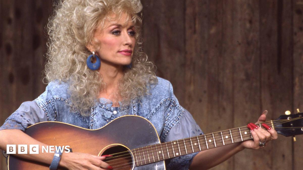 Dolly Parton strums a guitar while looking to the right of frame wearing a blue top against a background of wooden panels on ABC TV in 1987.