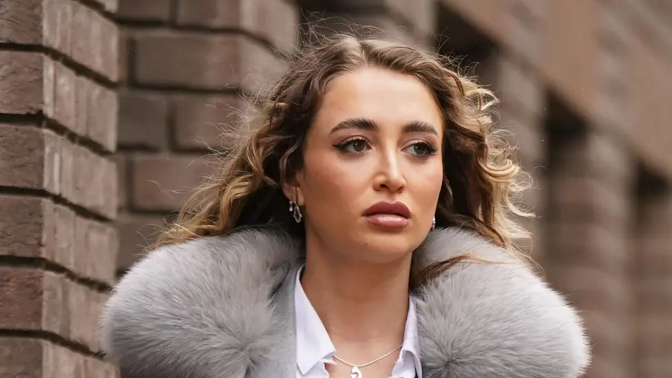 Georgia Harrison with long curled blonde hair, looking slightly away from the camera with a brick wall behind her. She is wearing a grey jacket and white shirt, and her overcoat has a big, fluffy grey collar.