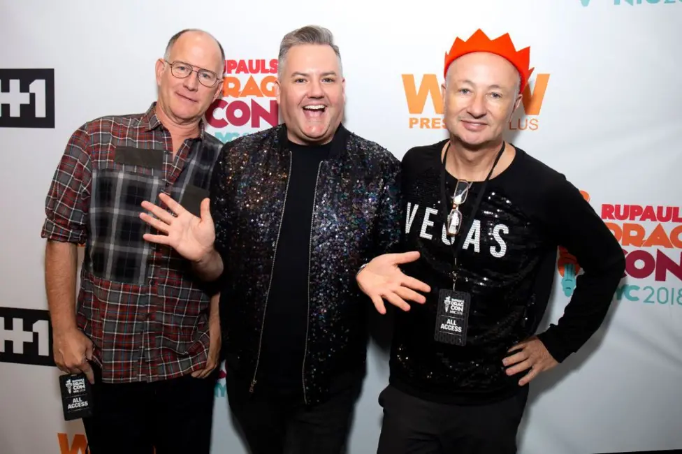 Three people standing in front of an event backdrop with visible logos and text including “RuPaul’s DragCon NYC 2018”, “WOW Presents Plus” and “#1”. The person in the centre wears a dark sequined jacket and raises both h