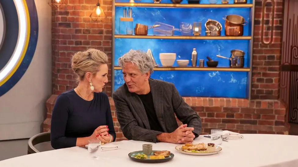 A woman with blonde hair tied up, wearing large earrings, a necklace chain, a bracelet and a dark blue long-sleeved top, with red nails is sat at a table with a man with grey hair, wearing a grey blazer and a black T-shi