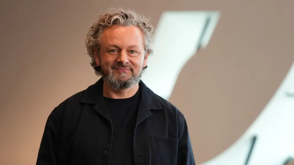 Michael Sheen who has curly, grey hair, a short beard and is wearing a dark shirt and jumper. He is smiling at the camera