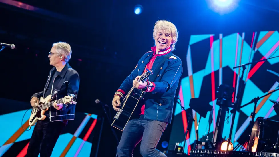 Jon Bon Jovi - and a bandmate to his right - play guitars on stage. They are both wearing jeans and blue jackets. Jon Bon Jovi has a big smile on his face and is wearing a red shirt. There is a drum kit in the background