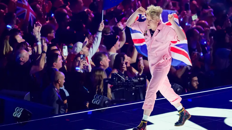 A performer wearing a pale pink one-piece outfit walks along a brightly lit Eurovision stage draped in a large Union Jack flag. Audience members line both sides of the stage, many holding phones and flags. Purple and blu