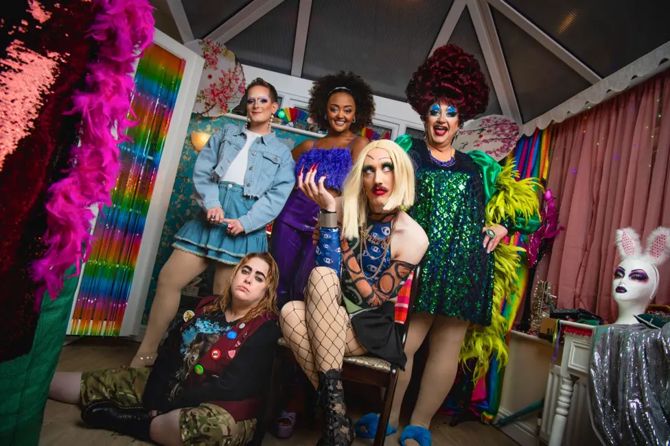 A promotional shot for Smoggie Queens which shows three men in drag posing in a conservatory alongside two women. In the foreground, one person sits on a chair wearing a blonde wig, fishnet tights, a patterned blue top a