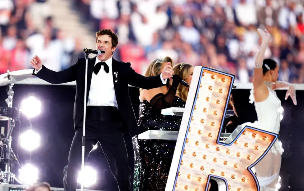 Brandon Flowers performs with The Killers at the UEFA Champions League final. He's dressed in a tuxedo with a bow tie, standing next to a large billboard-style letter K.