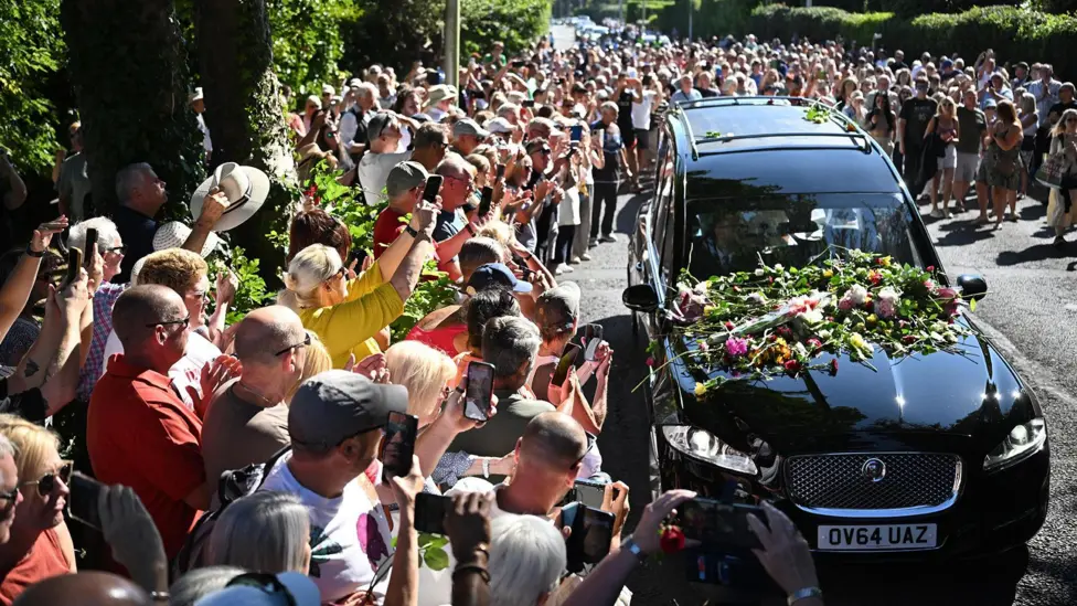 Bystanders flock towards a black hearse and throw flowers onto its bonnet as it drives through the town carrying the coffin of Bonnie Tyler.