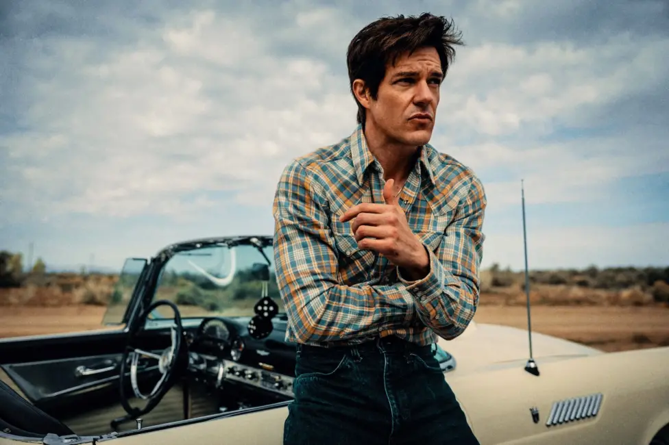 Brandon Flowers in a checked shirt, leaning against a classic American convertible, with fluffy dice hanging from the rear view mirror