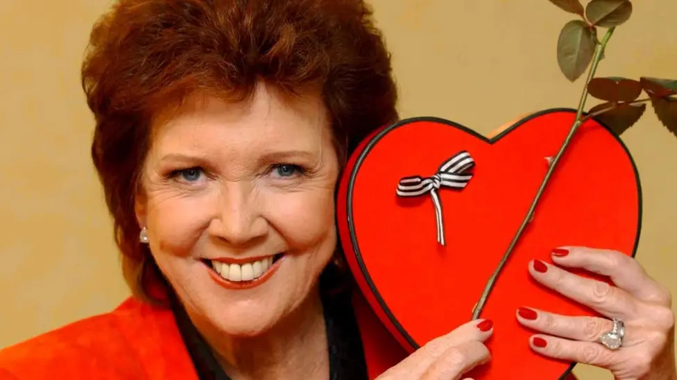 Cilla Black smiling and holding up a love heart while host of ITV show Blind Date