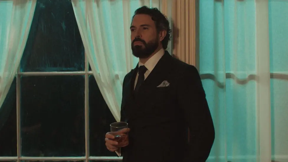 Tom wears a black suit with a white shirt and red tie, He has a grey pocket square.