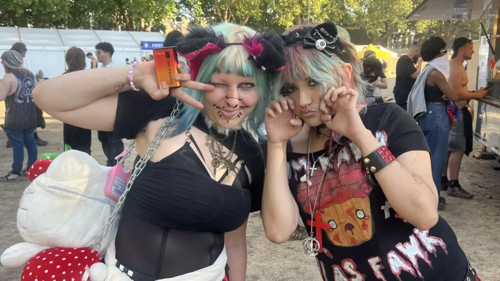 All Points East festival goers Bin and Zari smile at the camera wearing their festival gear. Bin wears a sheer black top and has green and blonde hair with a black furry headband. Zari wears a black slogan t-shirt and ha