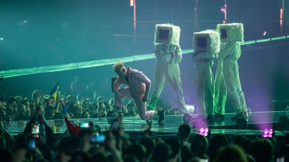 A performer in a pale pink outfit kneels on a Eurovision stage holding a microphone while singing. Behind are three large pale green furry characters with rectangular black screens instead of faces. The characters wear m