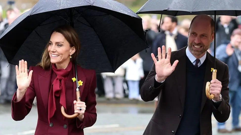 Catherine and William, Prince and Princess of Wales, on a trip to Wales. They are smiling and waving and wearing yellow daffodils on their coats