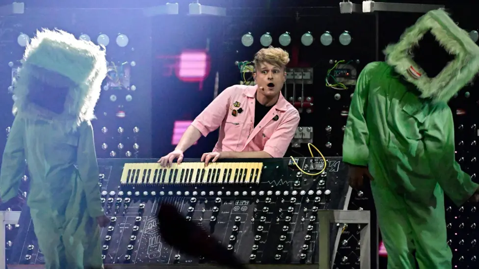 A performer in a pale pink outfit stands behind a large custom-built electronic keyboard and control console on a Eurovision stage. Two large pale green furry characters stand on either side of the instrument.