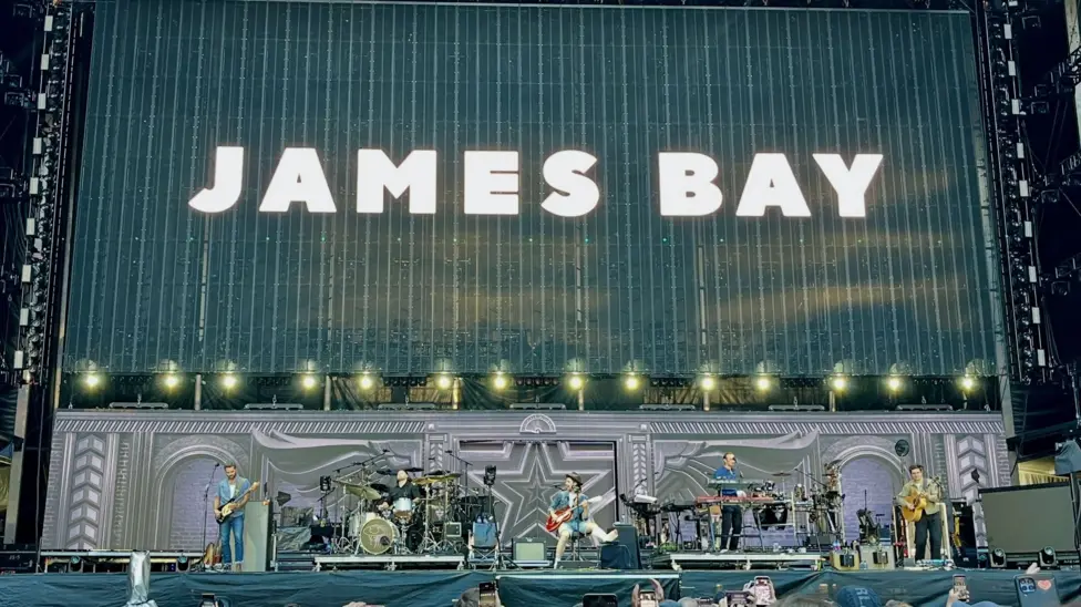 James Bay's name appears in white big letters on a black backdrop behind him and his band on stage.