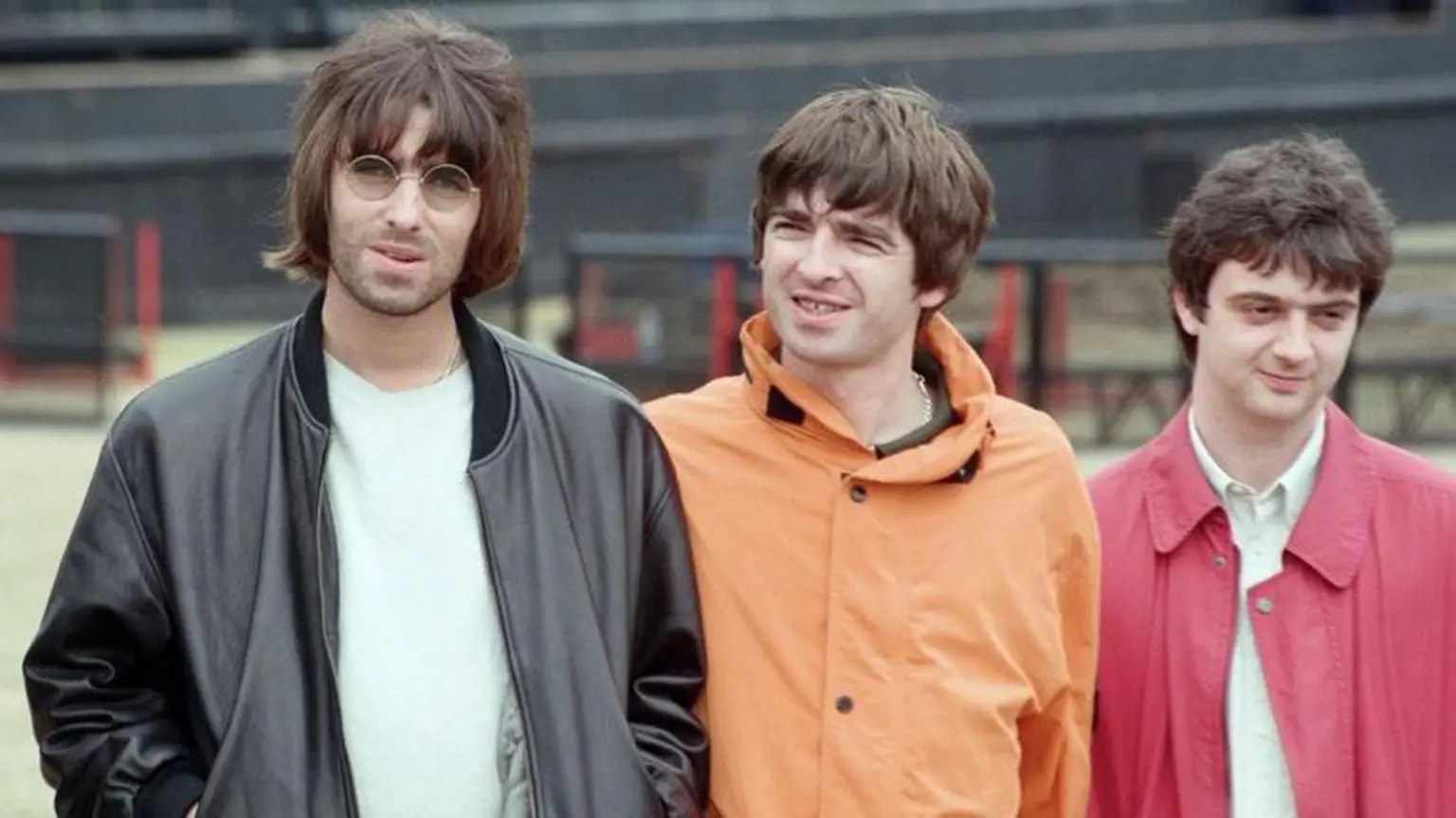 The Gallagher brothers together with their band at Knebworth in 1996. Liam has on a black leather jacket and round sunglasses while Noel is wearing an orange cagoule