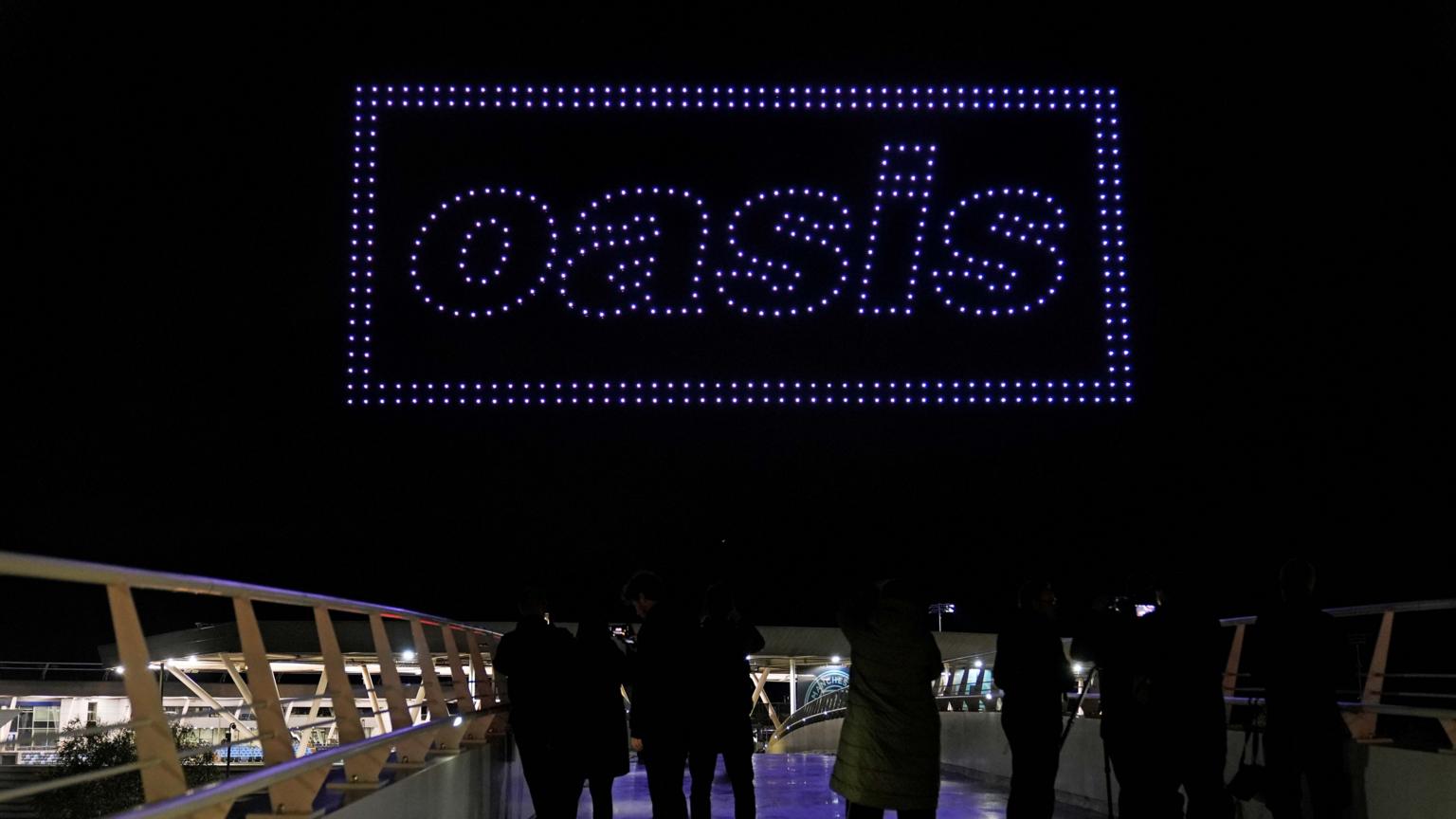 A drone light show teasing an Oasis tour announcement over a stadium skyline.