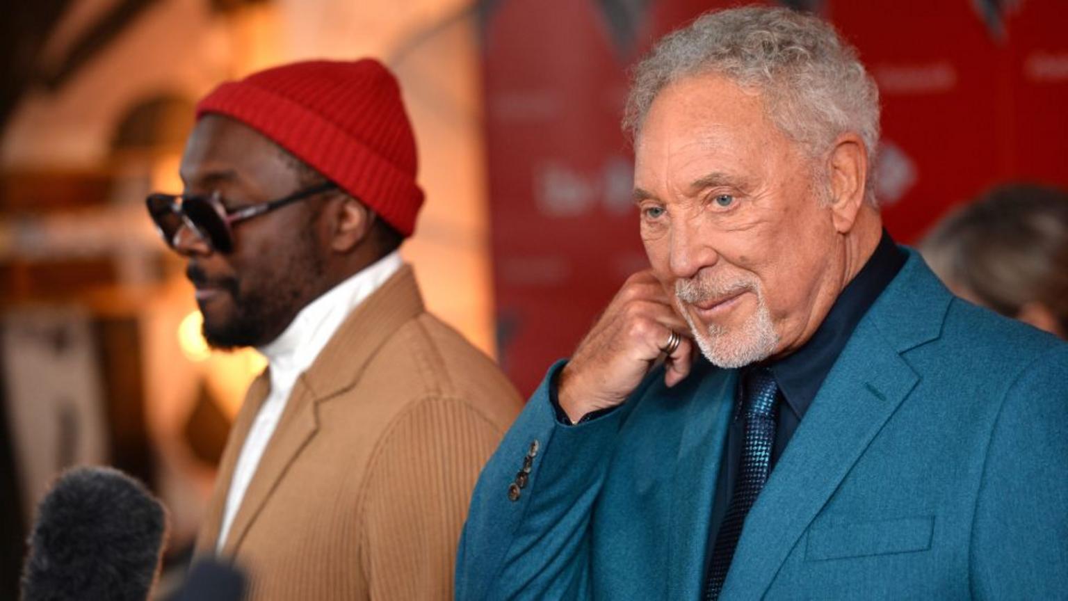 Sir Tom Jones, wearing a blue jacket, pictured being interviewed next to Will.I.Am, who is wearing a brown jacket, sunglasses and a red hat