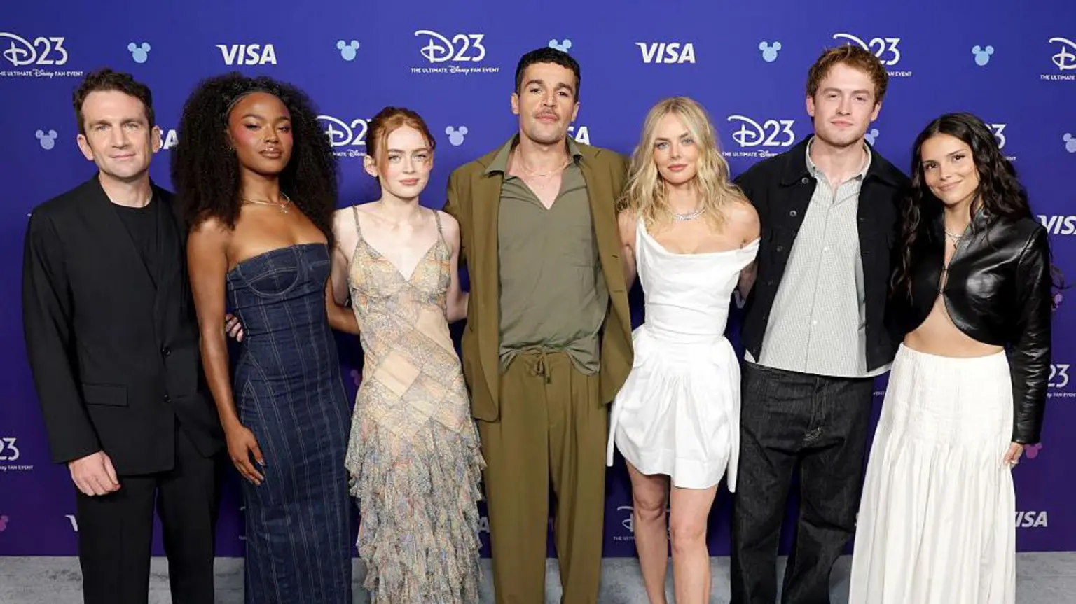 The cast of the new X-Men movie all standing up posing for a picture. Jake is in all black, Maya is in a dark blue dress, Sadie is wearing a dress with a rectangle pattern on it, Christopher is wearing an olive blazer an