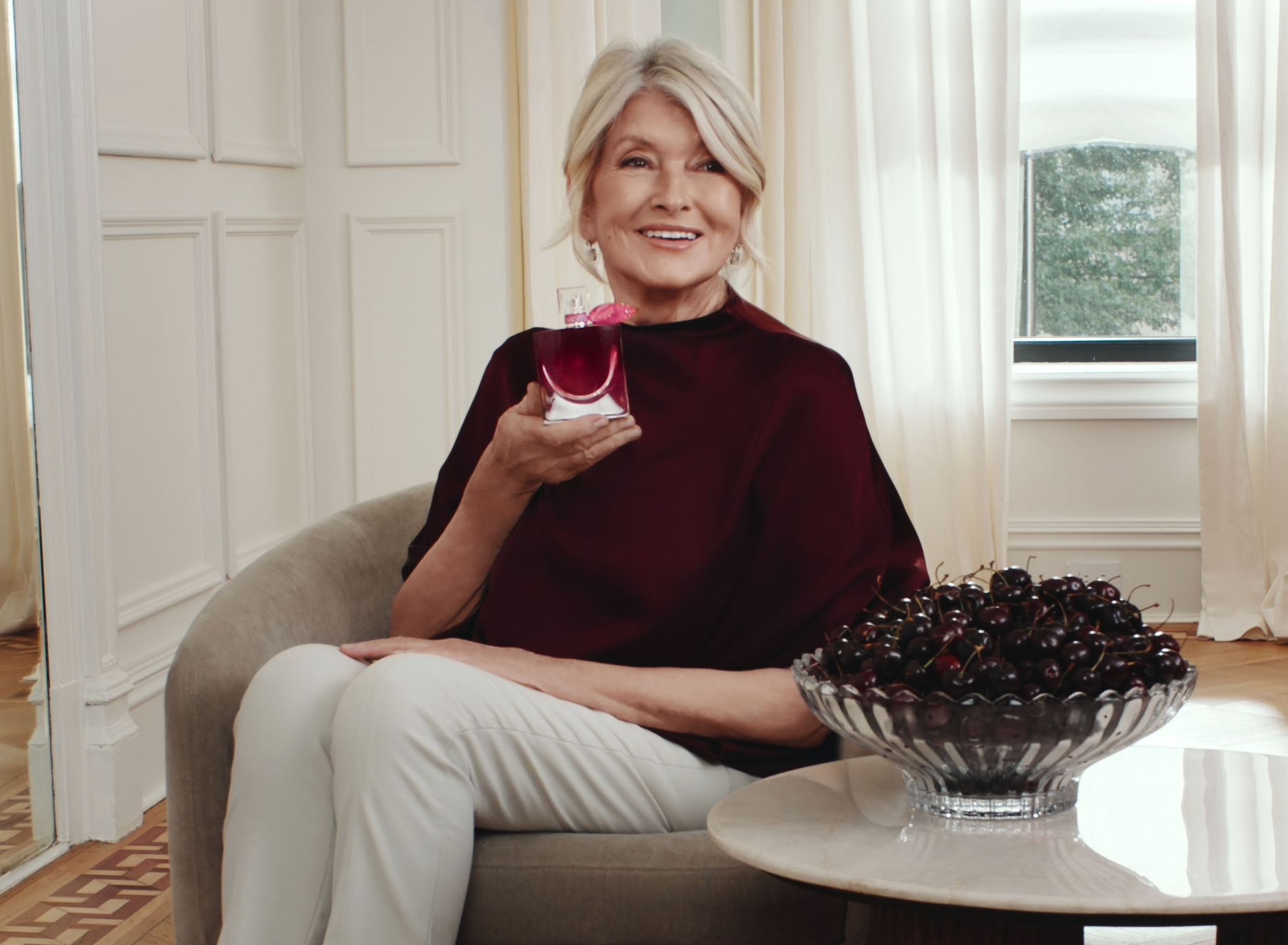 Martha Stewart seated, holding a bottle of perfume, with a bowl of cherries next to her.
