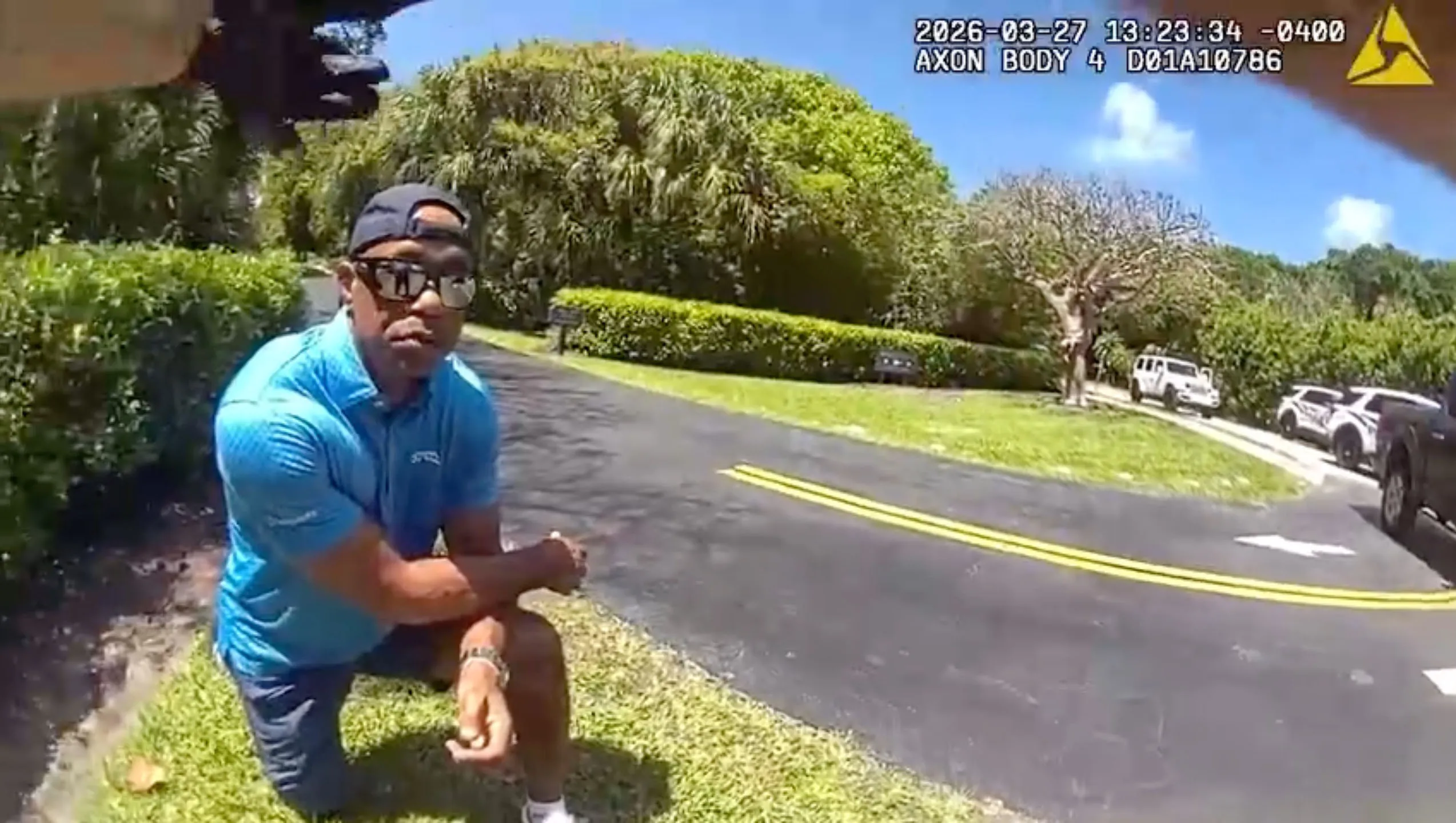 Bodycam footage showing a man in a blue shirt and hat kneeling on a lawn by a road, with police vehicles in the background.