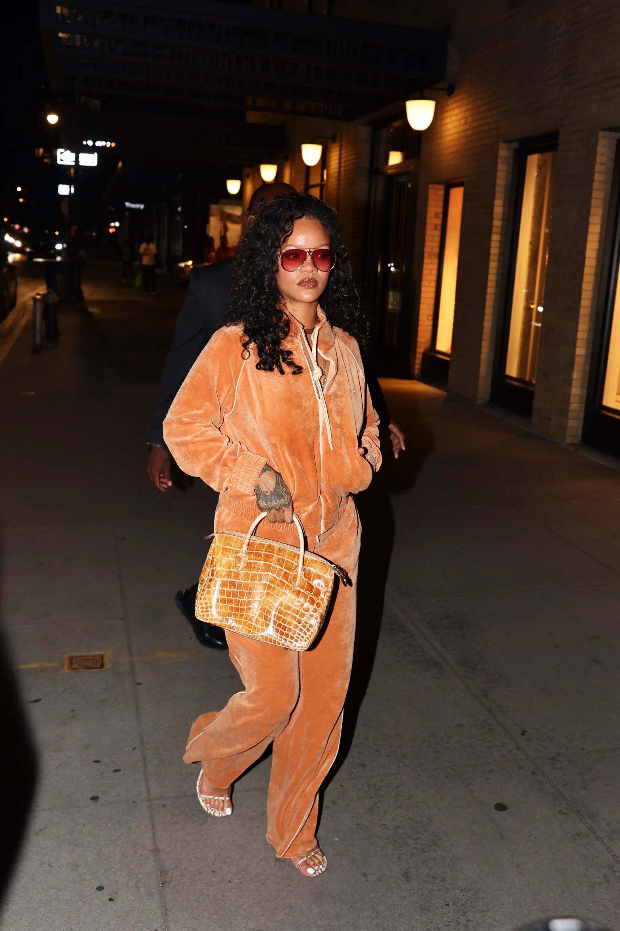 Rihanna walking on a city street at night in an orange velvet outfit and red-tinted sunglasses.