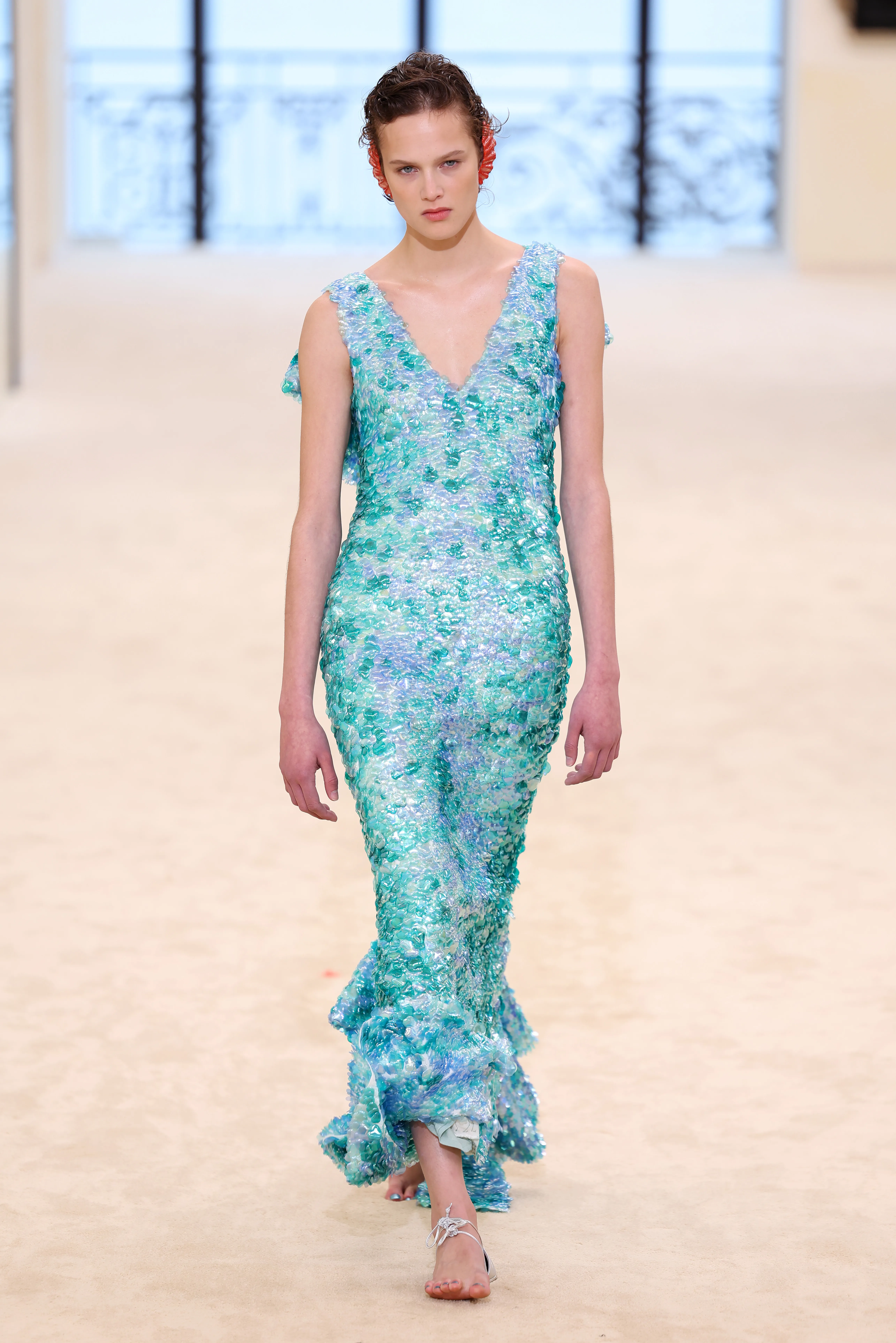 Noor Khan walks the runway in a blue and turquoise sequined dress with red ear pieces.