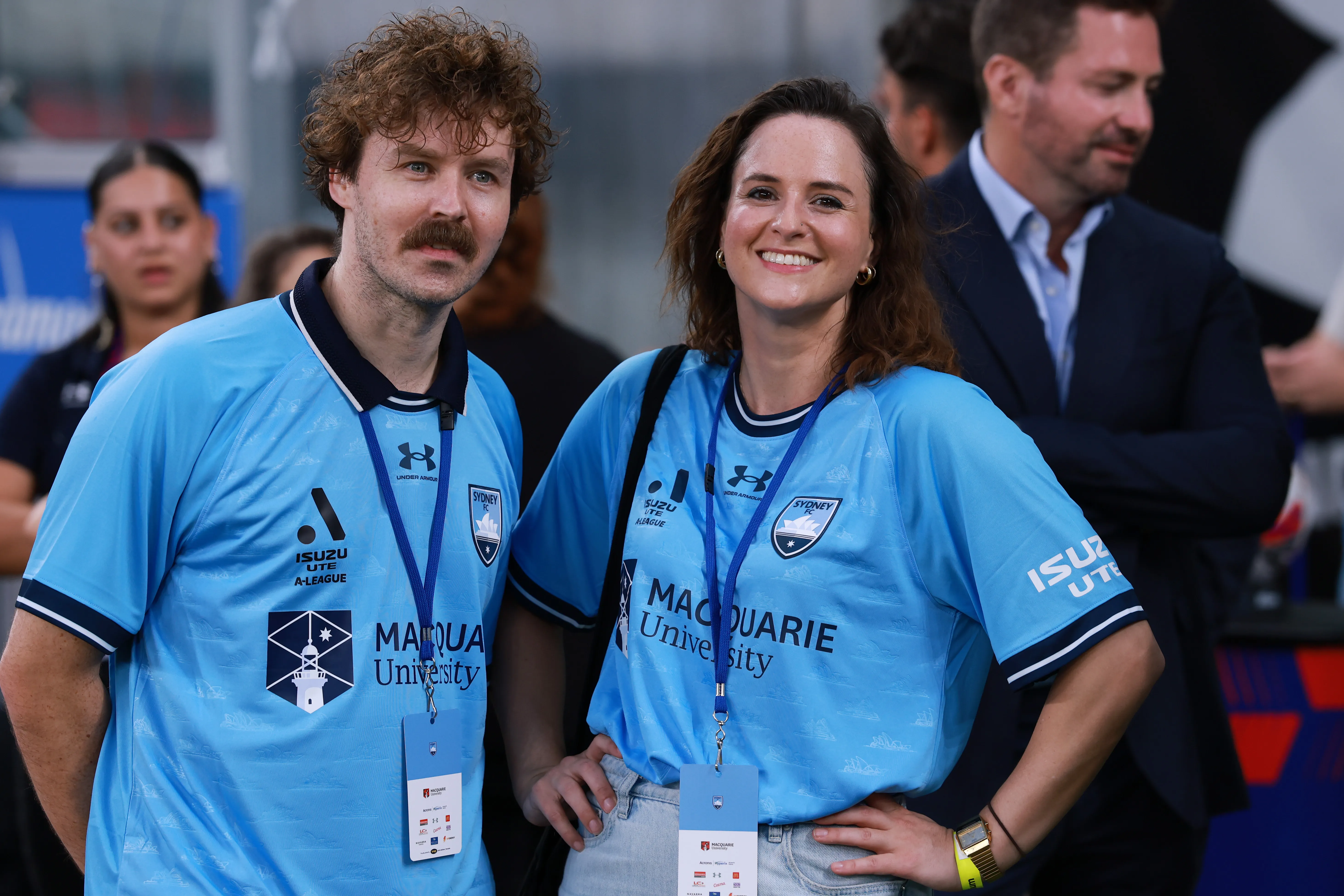 Australian Olympian Rachael Gunn, smiling, with a man, both wearing light blue Sydney FC shirts with sponsor logos.