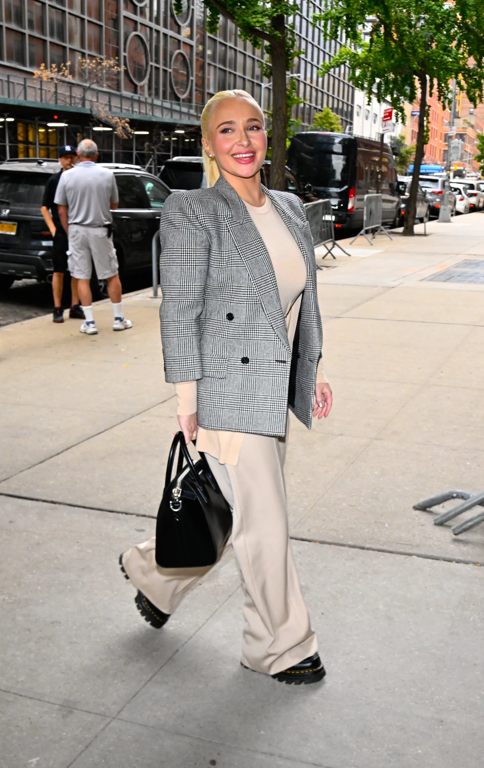 Hayden Panettiere wearing a plaid blazer, beige shirt and pants, and carrying a black handbag, walking down a New York City street.