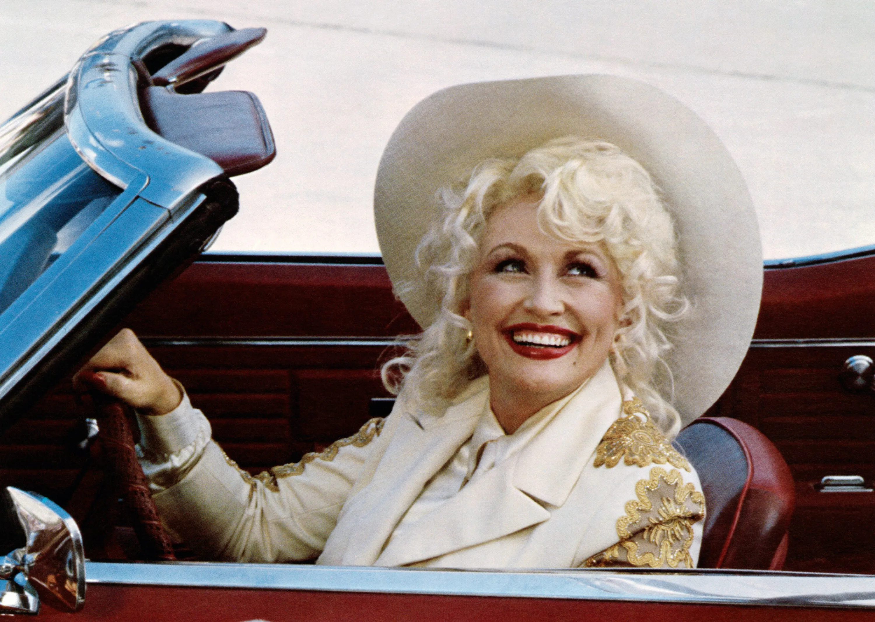 Dolly Parton in a convertible, wearing a white cowboy hat and a white jacket with gold embroidery.