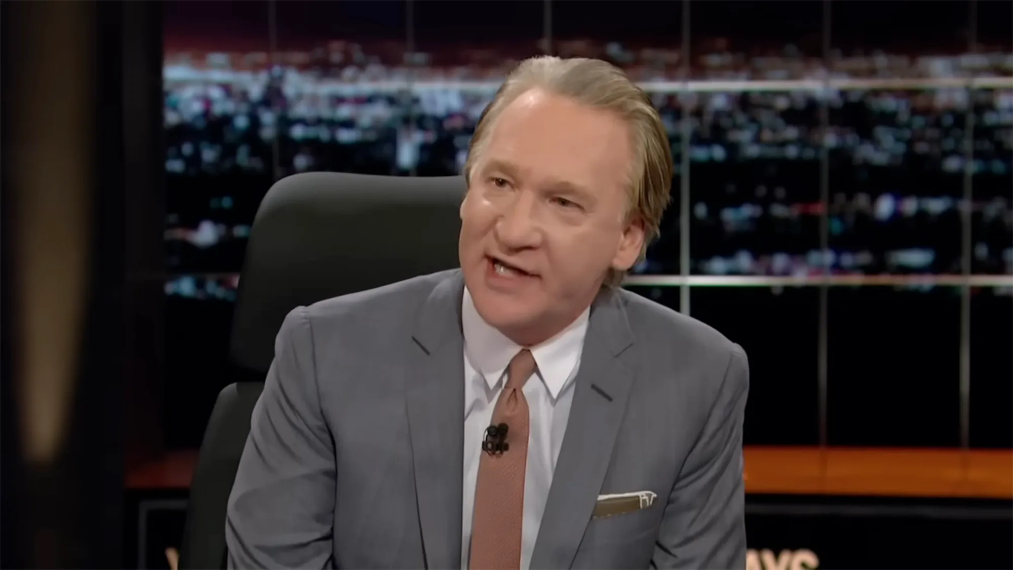 Bill Maher speaking with his mouth slightly open, wearing a gray suit and a salmon-colored tie.