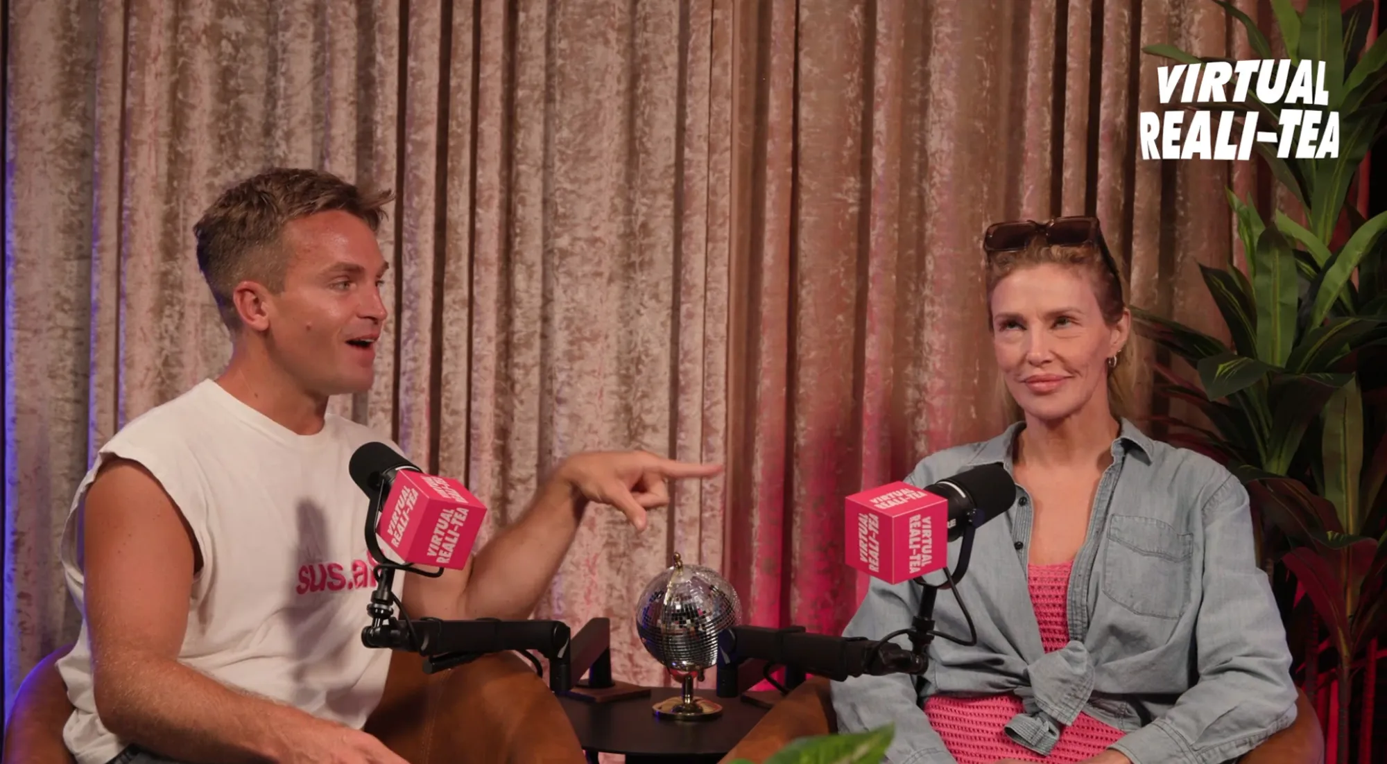 A man in a white t-shirt pointing to a woman in a denim shirt over a pink top, both with microphones in front of them labeled "VIRTUAL REALI-TEA".