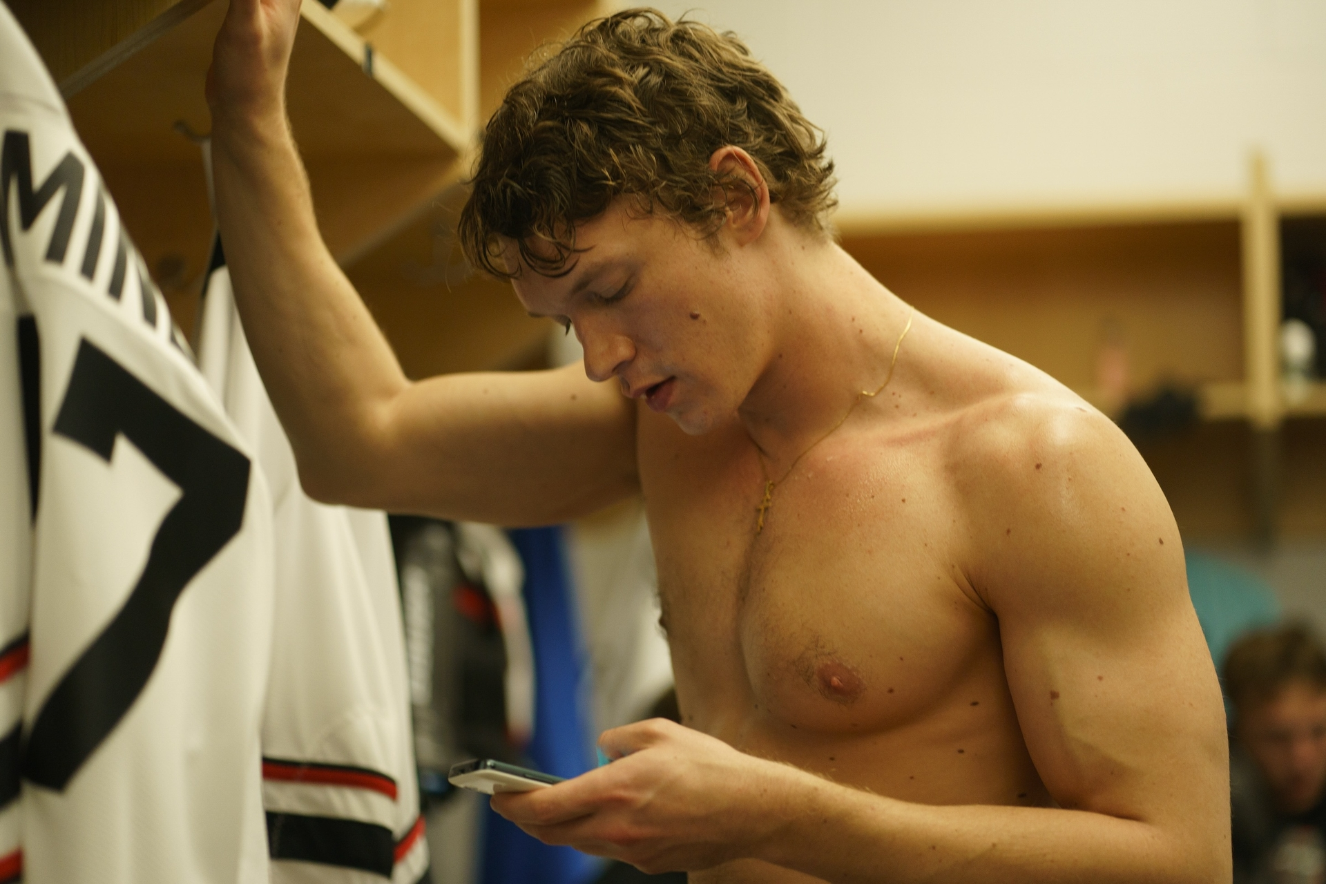 Connor Storrie as Ilya Rozanov in Heated Rivalry, shirtless and looking at a phone in a locker room.