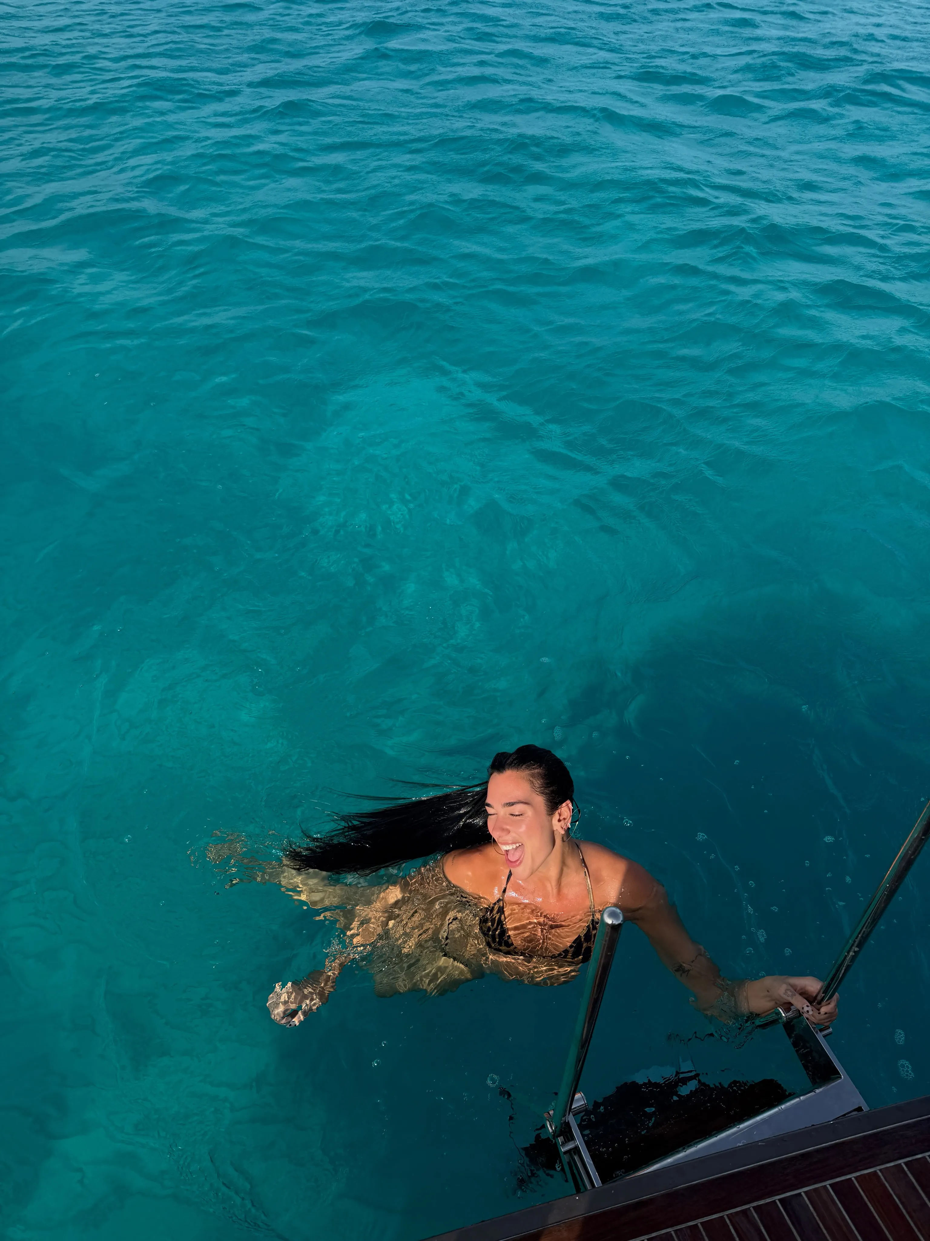 Dua Lipa in a black bikini swimming in blue-green water, grabbing onto a boat ladder.