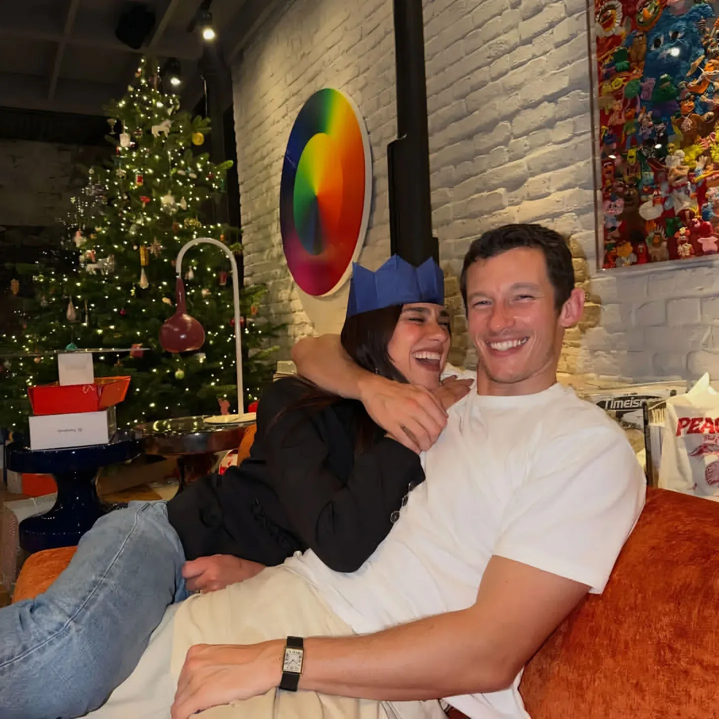Dua Lipa and Callum Turner embracing on a sofa in front of a Christmas tree.