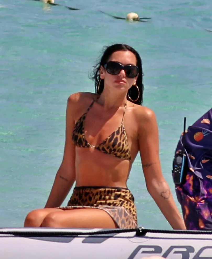 Dua Lipa on a boat, wearing a leopard print bikini, sunglasses, and hoop earrings.