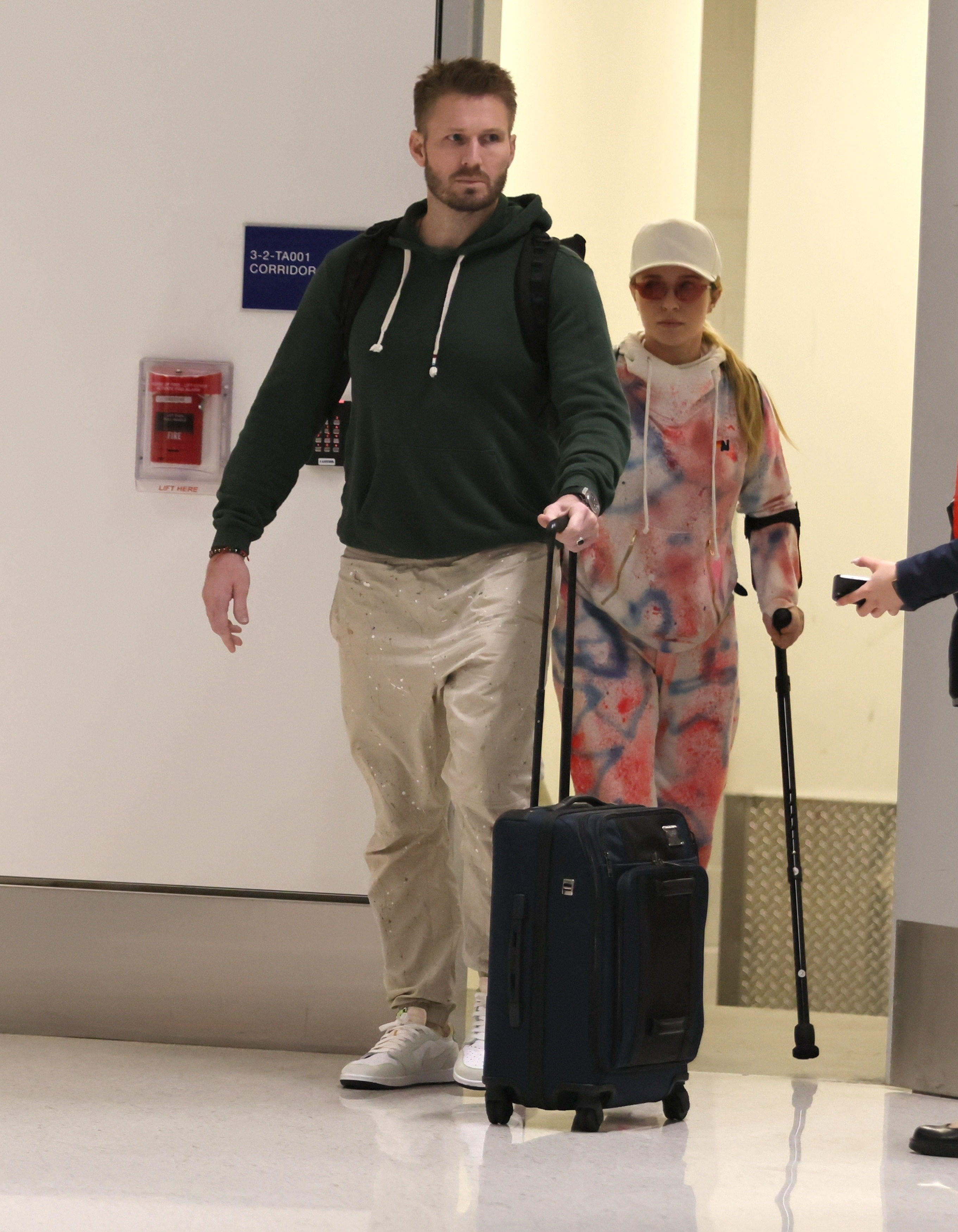 Hayden Panettiere on crutches and Brian Hickerson with a rolling suitcase walking in an airport.