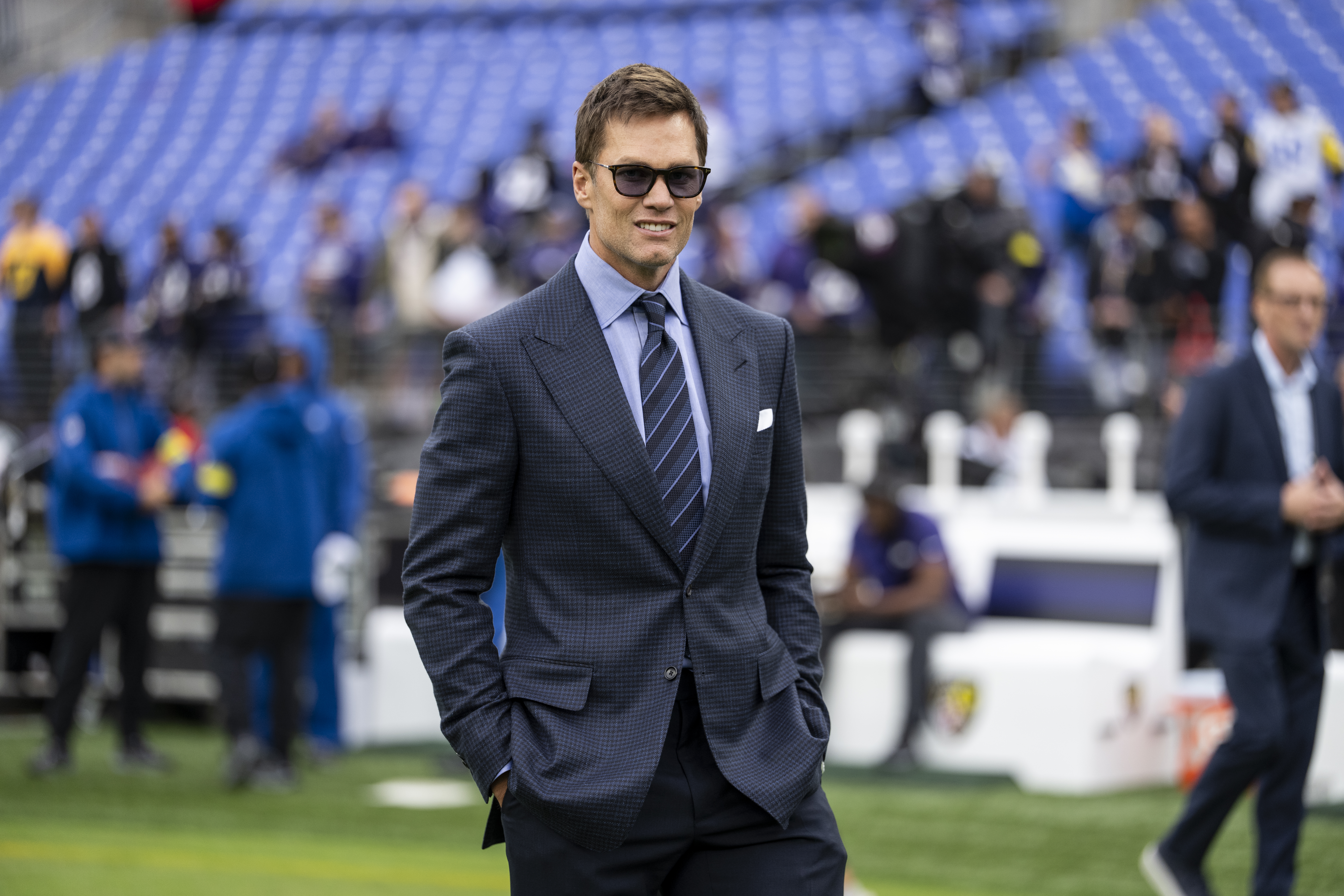 Former NFL quarterback Tom Brady wearing a suit and sunglasses on the sidelines of an NFL game.