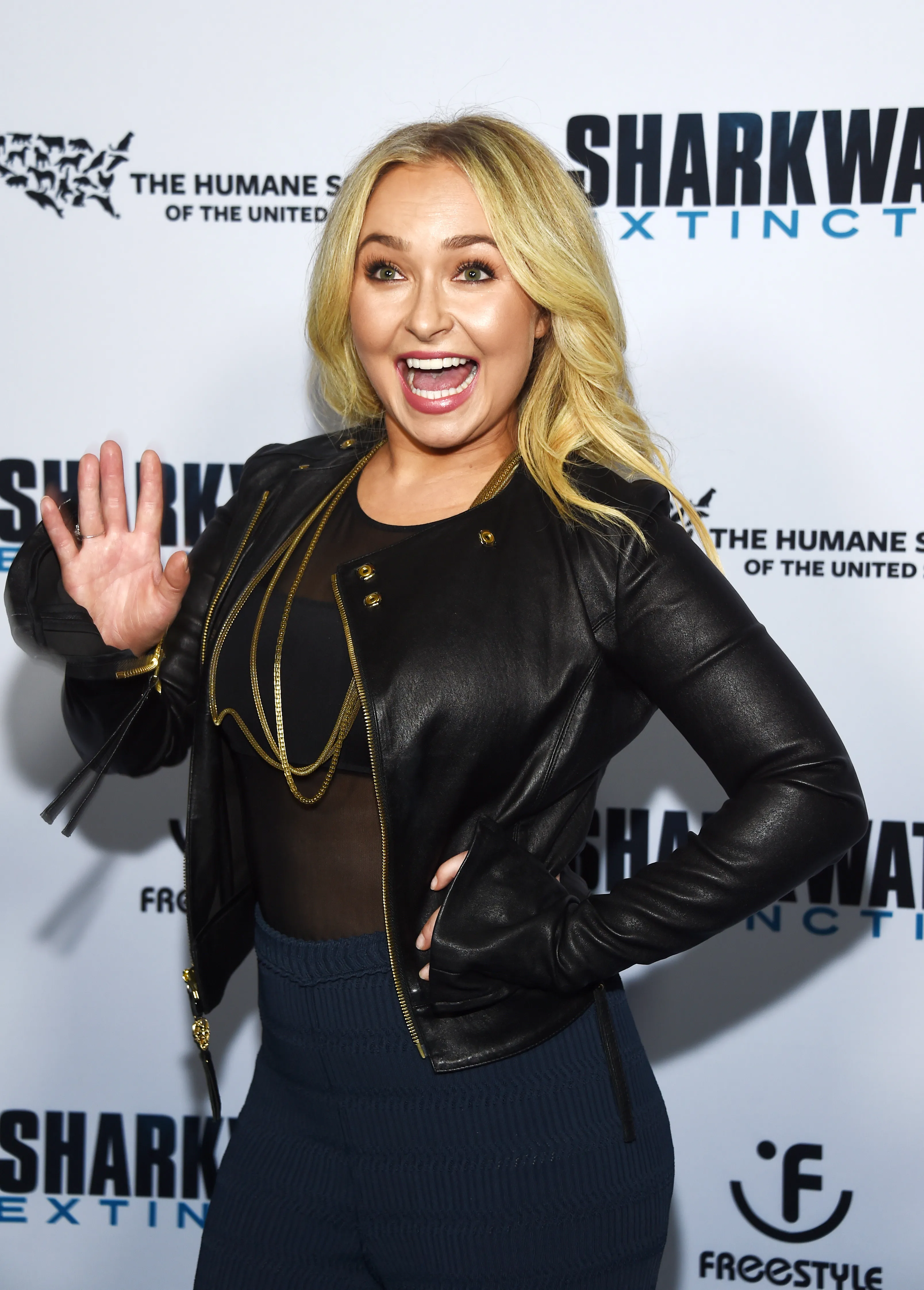 Hayden Panettiere in a black leather jacket and dark blue pants, with a "Sharkwater Extinction" logo behind her.