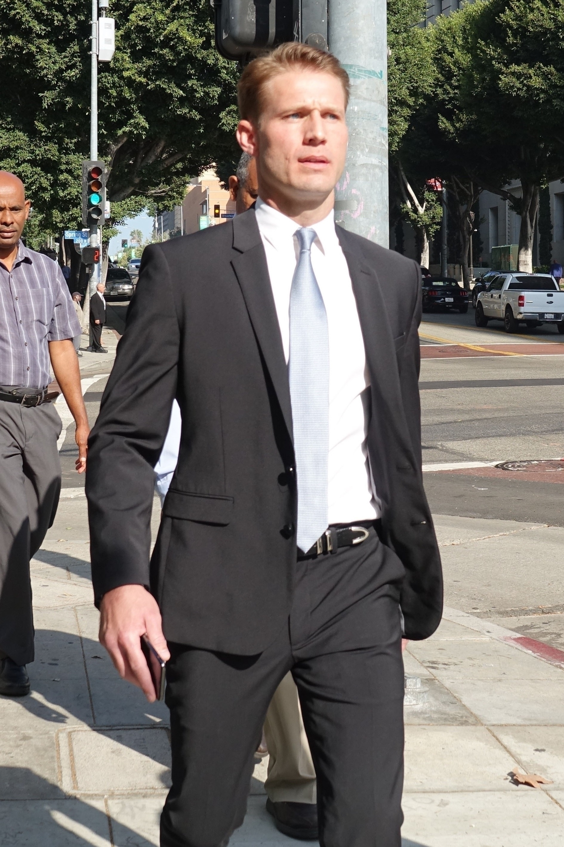 Brian Hickerson in a black suit with a light blue tie, walking on a city street.