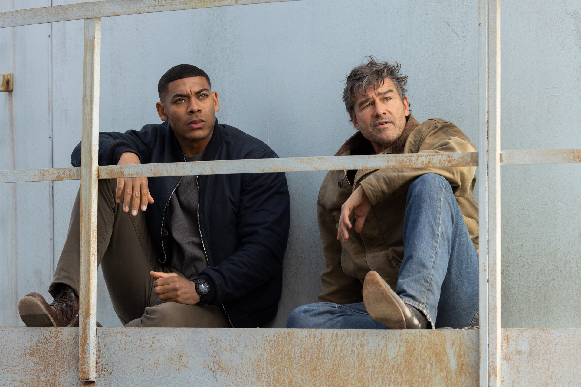 Two men, one Black and one white, sitting behind a rusty metal railing.
