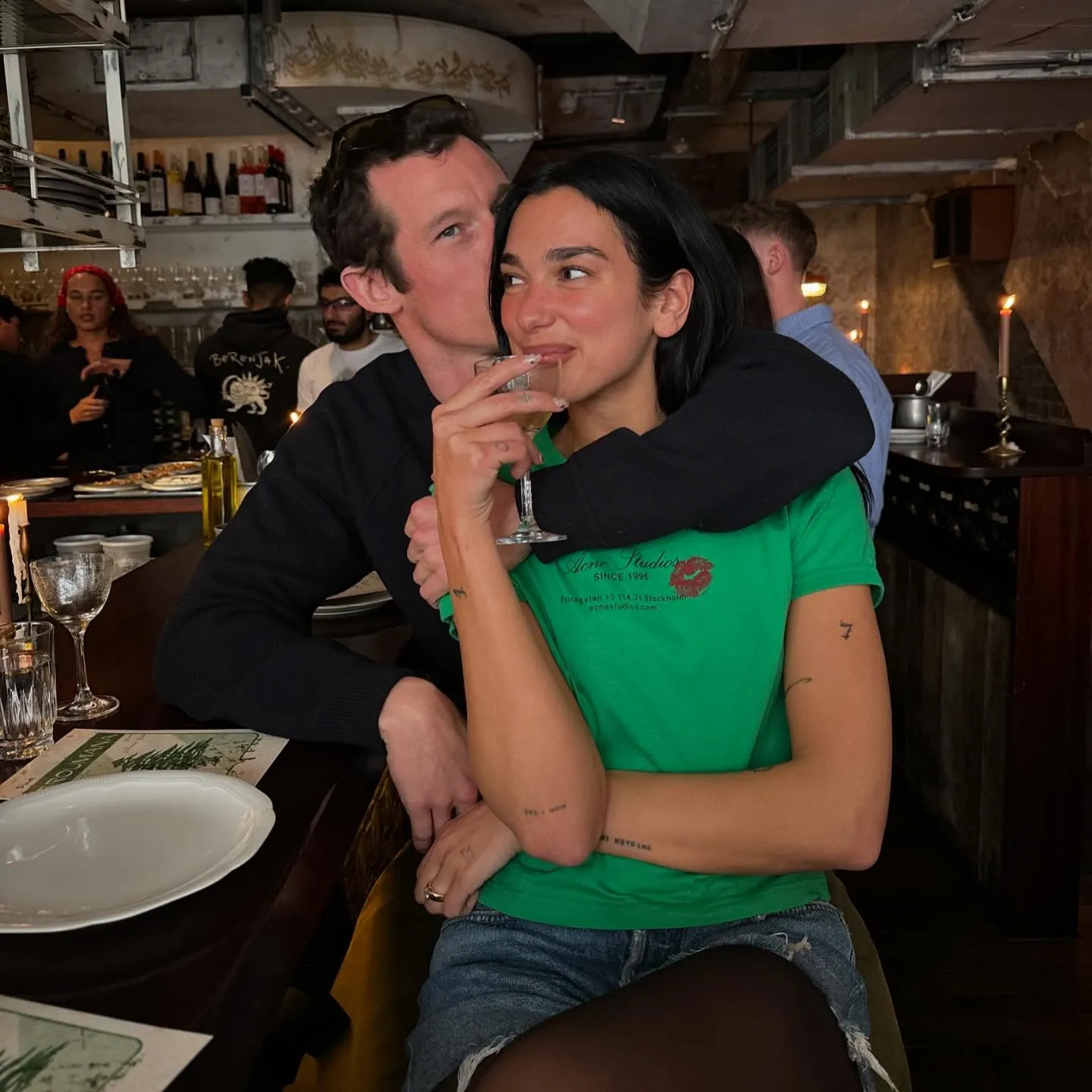 Dua Lipa smiles while Callum Turner kisses her cheek at a restaurant.